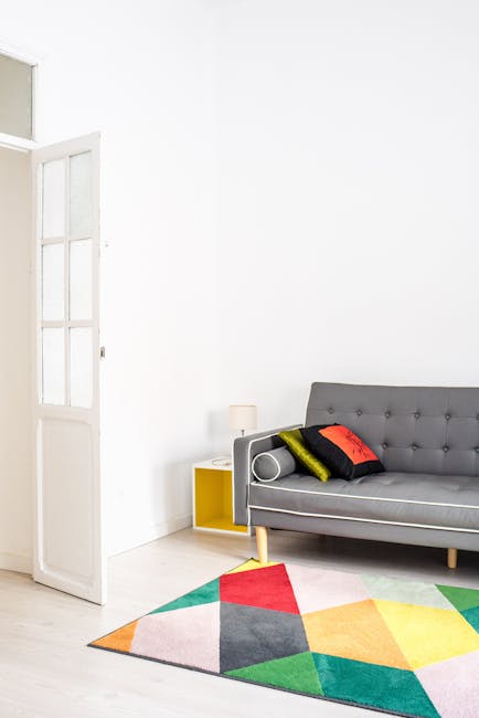 A bright, clean living room featuring a grey fabric sofa with button-tufted detailing and four decorative cushions in black, yellow, red, and light grey. The room has white walls and a white wooden floor, with natural light streaming through an open glass-paned door on the left. A modern, multicolored geometric area rug with shades of red, yellow, green, blue, pink, and grey lies partially beneath the sofa. A small yellow side table with a white lamp is next to the sofa, creating a tidy and well-maintained space. The scene showcases a neat and hygienic environment, indicative of professional domestic cleaning from Carpet Cleaning SE19, specializing in surface cleaning and deep cleaning services in the Crystal Palace Park area, SE19.