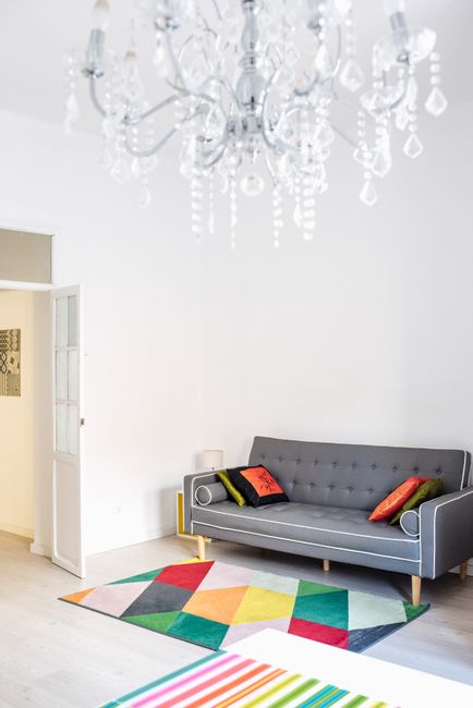 A bright, modern living room with white walls and light wooden flooring. A grey fabric sofa with a sleek, contemporary design is positioned against the wall, decorated with colorful cushions in red, orange, and green. In front of the sofa, a geometric-patterned area rug with vibrant hues of yellow, blue, red, and green covers part of the floor, adding visual interest and warmth to the space. To the left, a white door with glass panels is slightly open, revealing a glimpse into another room. A large, elegant chandelier with multiple tiers of hanging crystal droplets is suspended from the ceiling, providing ambient lighting and adding a touch of sophistication. The room appears clean and well-maintained, with no visible dust or clutter, exemplifying professional domestic surface cleaning and ongoing maintenance, perfectly aligned with the services of Carpet Cleaning SE19 for same-day carpet cleaning in the Crystal Palace Park area, SE19.