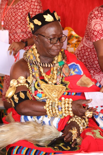 Close-up of a woman wearing traditional African attire with intricate gold jewelry, including necklaces, bracelets, and a headpiece, sitting on a red household furniture piece. The background features